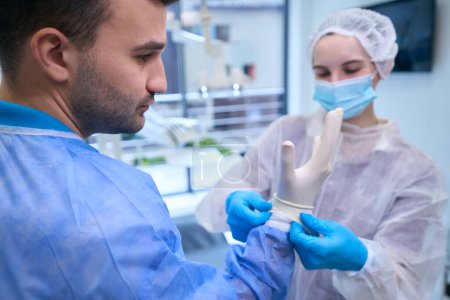 Téléchargez les photos : Infirmière met sur le médecin des gants stériles jetables, l'équipe se prépare pour l'opération - en image libre de droit
