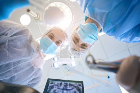 Zahnarzt Chirurg mit spezieller Pinzette und herausgezogenem Zahn in den Händen im Operationssaal, ihm hilft eine junge Assistentin