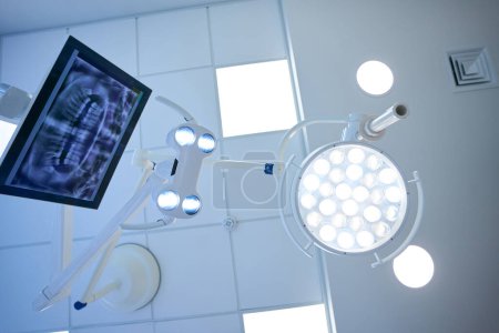 Téléchargez les photos : Lampe chirurgicale et moniteur avec l'image d'une radiographie dentaire sur le lieu de travail d'un dentiste dans une clinique moderne - en image libre de droit