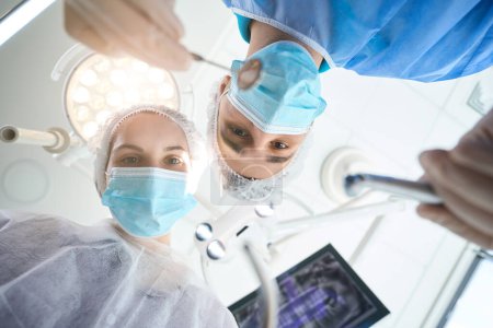 Téléchargez les photos : Chirurgien avec un miroir dentaire dans les mains sur le lieu de travail dans la salle d'opération, une assistante l'aide - en image libre de droit