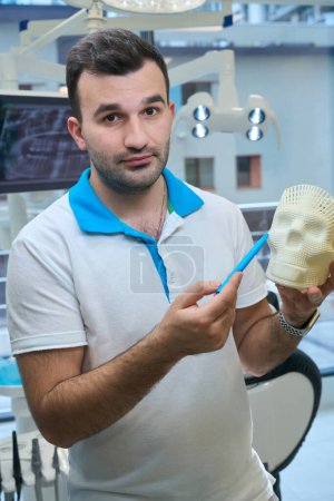Foto de Hombre en el lugar de trabajo en la silla dental, tiene una maqueta de un cráneo humano en sus manos - Imagen libre de derechos
