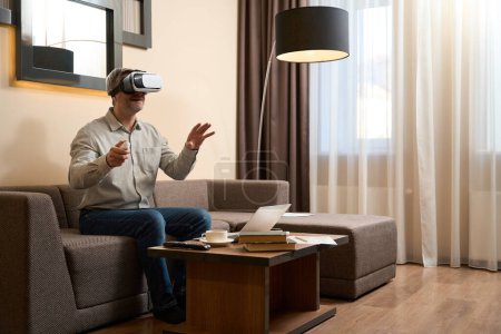 Foto de Hombre explorando gafas de realidad virtual y sosteniendo el controlador mientras está sentado en el sofá en la sala de hotel - Imagen libre de derechos