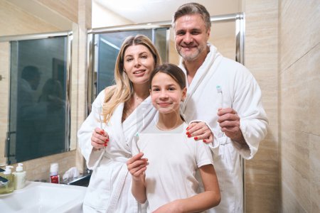 Photo for Happy young girl, man and woman in white bathrobe holding toothbrushes and ready to brush teeth - Royalty Free Image