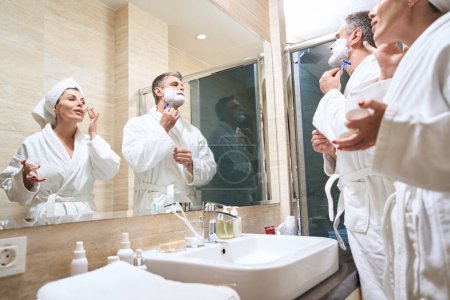 Téléchargez les photos : Homme et femme en manteaux blancs debout près du miroir, mari se rase, femme applique de la crème sur la peau dans l'hôtel - en image libre de droit