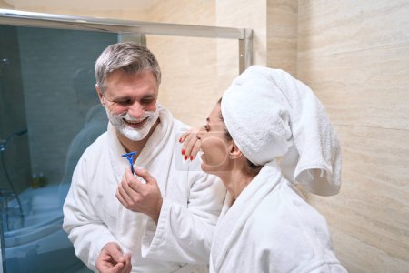 Foto de Hombre y mujer adultos con abrigos blancos mirándose, el marido se afeita en la habitación del hotel - Imagen libre de derechos