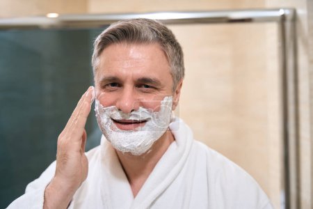 Téléchargez les photos : Homme adulte en manteau blanc applique de la mousse sur son visage dans la salle de bain, en regardant la caméra - en image libre de droit
