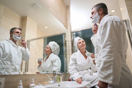 Téléchargez les photos : Homme en manteau blanc debout à côté de sa femme et prendre soin de son visage, femme buvant du café dans la salle de bain - en image libre de droit