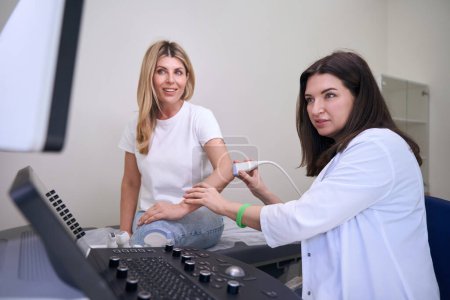 Foto de Diagnóstico médico lleva a cabo un examen por ultrasonido de la articulación del codo, las mujeres miran el monitor del dispositivo - Imagen libre de derechos