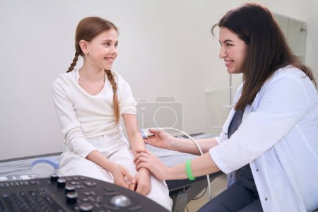 Foto de Amistosa mujer médico realiza un examen de ultrasonido de un paciente joven, el médico trabaja con la articulación del codo - Imagen libre de derechos