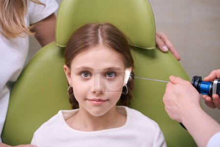 Foto de Chica bonita está sentado en un sillón verde en la cita con un otorrinolaringólogo, el médico examina el oído de los niños - Imagen libre de derechos