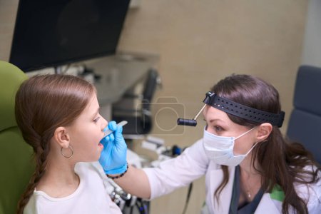 Foto de Médico en guantes protectores y una máscara examina la garganta de un niño, un otorrinolaringólogo utiliza herramientas especiales - Imagen libre de derechos