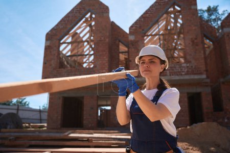 Photo for Concentrated woman carpenter in uniform and hardhat choosing proper and straight wooden beam, working at roof building at construction site - Royalty Free Image