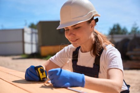 Photo for Close-up woman building engineer working with wooden beams, measuring necessary size with tape measure and making notes on beams - Royalty Free Image