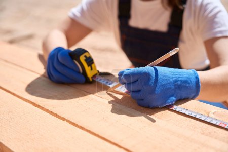 Photo for High-qulified carpenter measuring needed size with tape measure and making notes on wooden beams with pencil, close-up - Royalty Free Image