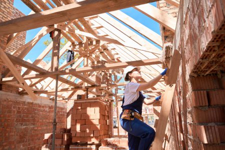 Photo for Confident woman building engineer climbing wooden ladder up to second floor of building under construction, looking at wooden roof frame construction - Royalty Free Image