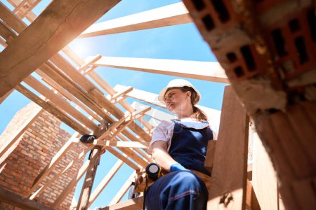 Photo for Bottom view responsible for roof building woman standing on ladder and checking quality and readiness of wooden roof frame, construction site - Royalty Free Image