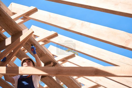 Foto de Mujer atenta contratista general en hardhat martillar un clavo en el marco de madera de la construcción del techo, trabajador en el sitio de construcción - Imagen libre de derechos