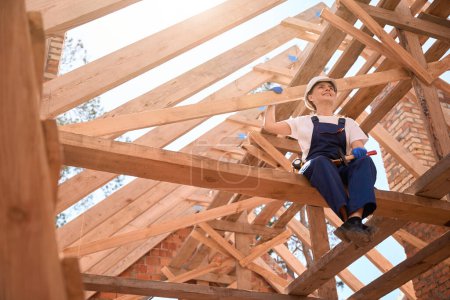 Photo for Smiling woman roofer in uniform holding hammer, resting from ceiling work sitting on roof girder, waiting for materials, construction site works - Royalty Free Image