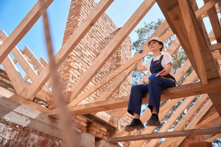 Photo for Responsible woman roofer in uniform and with hammer, sitting on roof beam and enjoying done roofing works, resting after hard work at construction site - Royalty Free Image