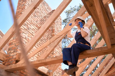 Photo for Cheerful woman roofer sitting on wooden beam of roof construction and talking phone, chatting with friends during break at construction site - Royalty Free Image