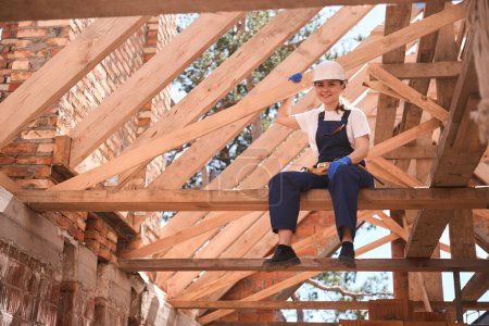 Foto de Alegre techador mujer de alta cualificación sentado en viga de madera y mirando el sitio de construcción, satisfecho con el trabajo hecho y buenos materiales - Imagen libre de derechos