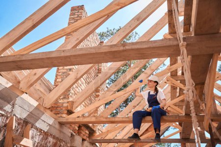 Photo for Responsible woman general contractor sitting on beam of roof frame and looking at construction site, satisfied with qualitative work - Royalty Free Image