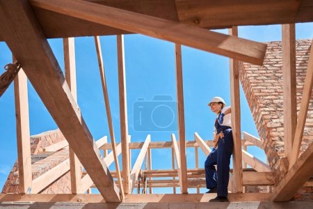 Photo for Forewoman in hardhat and uniform climbing up on wooden roof frame to look at construction site from top, enjoying with building progress - Royalty Free Image