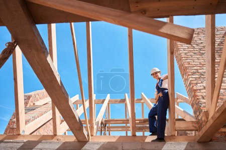 Photo for Smiling woman building engineer or roofer standing on wooden beam of roof frame and inspecting done job, enjoying view to construction site - Royalty Free Image