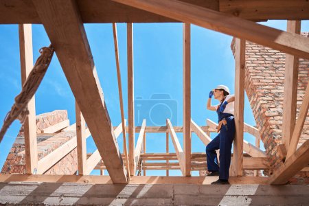 Photo for Skilled woman roofer adjusting hardhat standing on wooden frame of roof, resting after hard work and enjoying well done job, work at construction site - Royalty Free Image