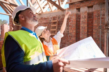 Photo for Woman architect showing somethimg on roof beams to serious foreman with blueprint, construction workers discussing building - Royalty Free Image