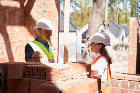 Photo for Male contractor talking to female architect near pile of bricks, workers in hardhats and hi vis safety vests discussing construction site - Royalty Free Image