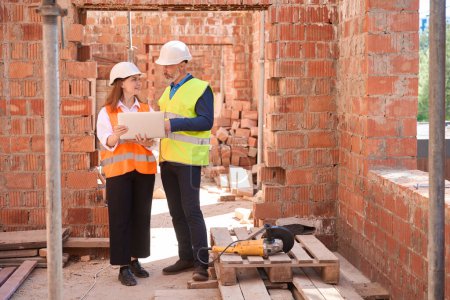 Photo for Female architect showing blueprint on laptop to building engineer who working on construction site with grinder tool - Royalty Free Image