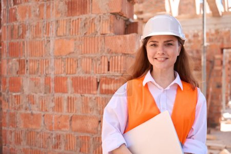 Photo for Positive woman architect in hardhat and orange safety vest holding laptop and smiling standing on construction site, 3d building modelling - Royalty Free Image