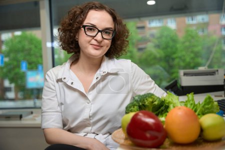 Foto de Mujer joven bonita en gafas se sienta en un espacio de oficina, al lado de un plato con verduras brillantes - Imagen libre de derechos