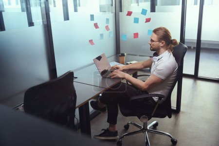 Téléchargez les photos : Homme avec des lunettes travaille dans un immeuble de bureaux sur un ordinateur portable, il est assis dans une chaise de bureau - en image libre de droit
