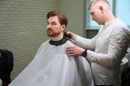Foto de Hombre guapo en peluquería peignoir sentado mientras peluquero diligente cortar su cabello mediante el uso de cortador eléctrico en el interior - Imagen libre de derechos