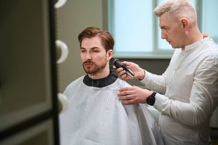 Photo for Diligent hairdresser making stylish haircut to his Caucasian client by using electric clipper in modern barbershop - Royalty Free Image