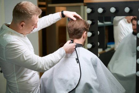 Photo for Skilled hair stylist trimming hair of his client wearing cutting hair cape in cool barber salon - Royalty Free Image