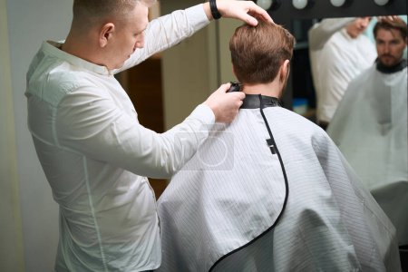 Photo for Handsome man looking at himself in mirror while hairdresser making trendy haircut for him indoors - Royalty Free Image