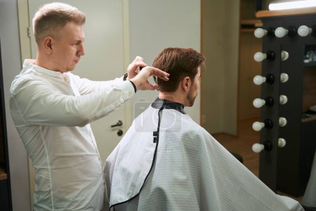 Foto de Peluquero calificado haciendo corte de pelo elegante a su cliente con tijeras y peine en la habitación moderna - Imagen libre de derechos