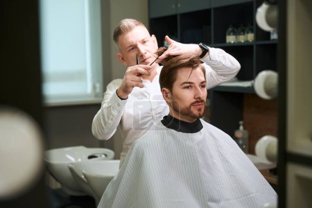 Photo for Professional barber diligently cutting hair of man by using hairdressing items in modern room - Royalty Free Image