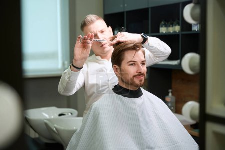 Photo for Handsome man with happy facial expression sitting while hairdresser cutting his hair by using scissors and comb indoors - Royalty Free Image