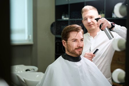 Foto de Peluquero experimentado mojar el cabello de su cliente mediante el uso de aerosol en la barbería de moda - Imagen libre de derechos