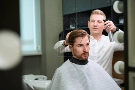 Téléchargez les photos : Barbier enthousiaste pulvérisation d'eau sur les cheveux du visiteur masculin dans la salle de salon moderne - en image libre de droit