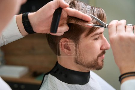 Foto de Vista recortada en las manos de peluquero recorte de pelo de hombre joven con tijeras en habitación moderna - Imagen libre de derechos