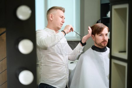 Photo for Caucasian barber providing services to his client who wearing cutting hair cape indoors - Royalty Free Image
