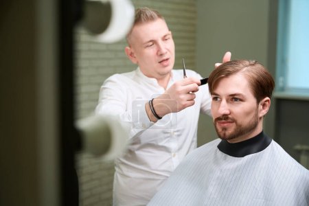 Photo for Skilled barber cutting hair of young man sitting in chair in modern barbershop - Royalty Free Image