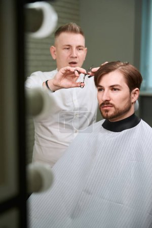 Photo for Young hairdresser cutting hair of his visitor by using scissors in modern barbershop - Royalty Free Image