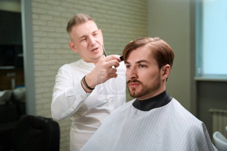 Photo for Handsome hairdresser making stylish haircut to his client who wearing in hairdressing peignoir indoors - Royalty Free Image