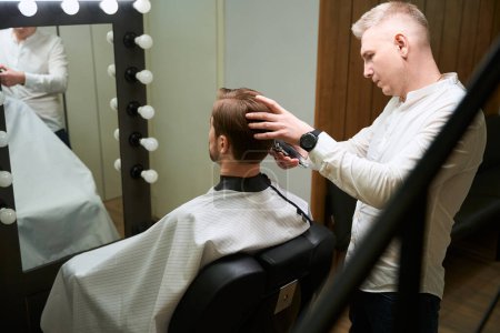 Photo for Caucasian hairdresser cutting hair of his client by using barber machine in modern salon - Royalty Free Image
